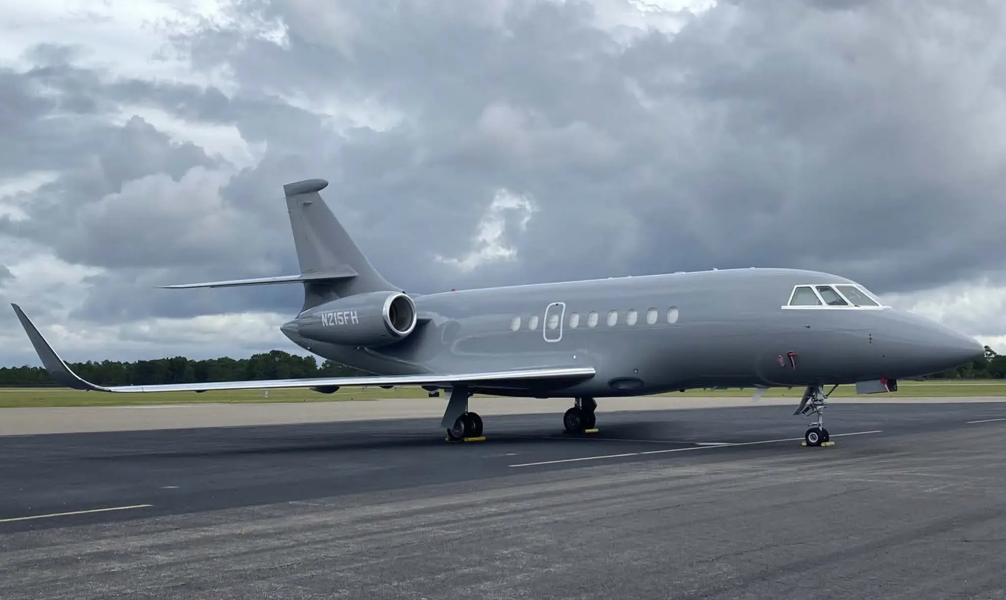 A Dassault Falcon 2000S business jet, registration N215FH, in a matte grey paint scheme, parked on an airfield under a cloudy sky.