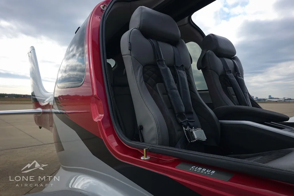 The gray leather front seats of a Cirrus SR22 aircraft, viewed through an open door on an airport apron.