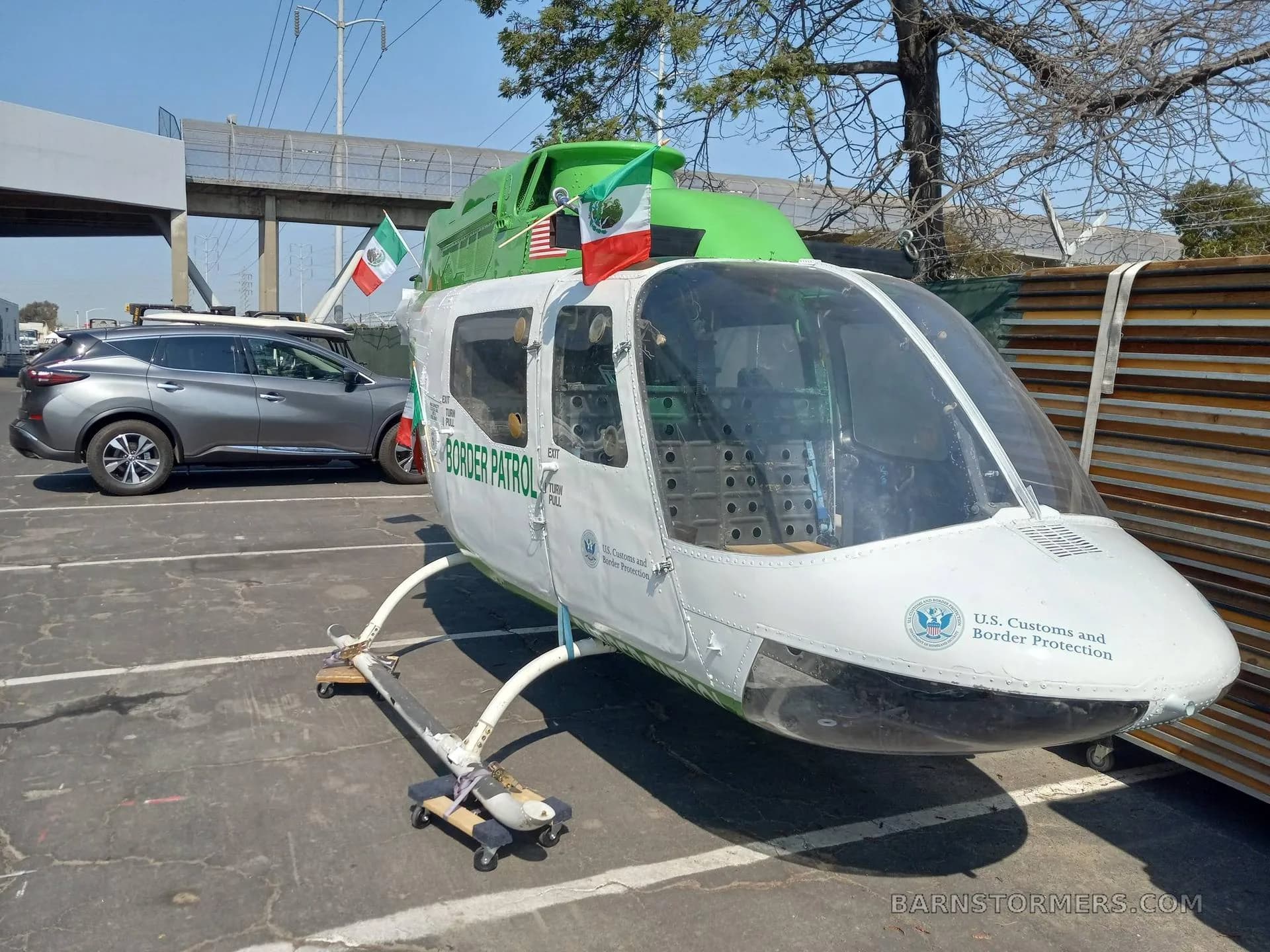 A white and green U.S. Customs and Border Protection helicopter fuselage, with Mexican flags, parked on asphalt next to a car.