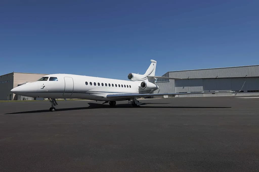 A white Dassault Falcon business jet is parked on an asphalt tarmac under a clear blue sky, with hangars in the background.