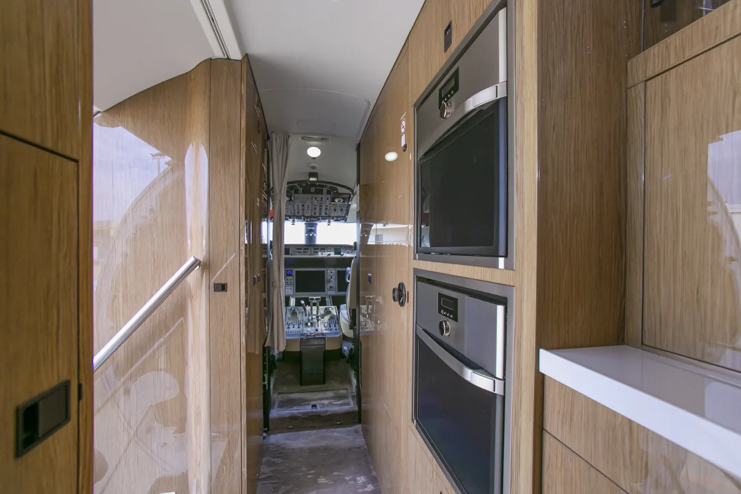 An aircraft galley with wooden cabinetry, built-in ovens, and a view towards the cockpit.