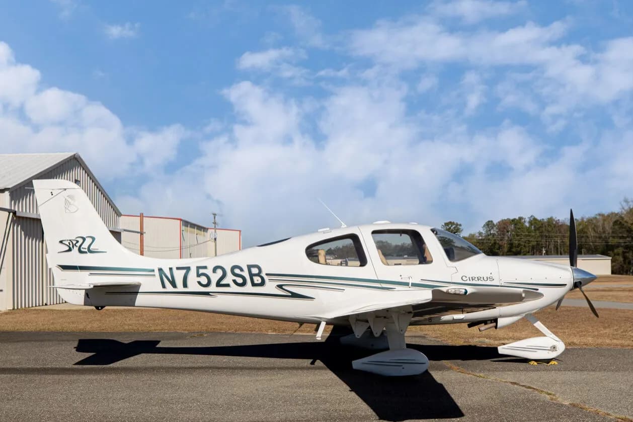 A side view of the white Cirrus SR22 aircraft, registration N752SB, parked on an asphalt ramp with trees in the background.
