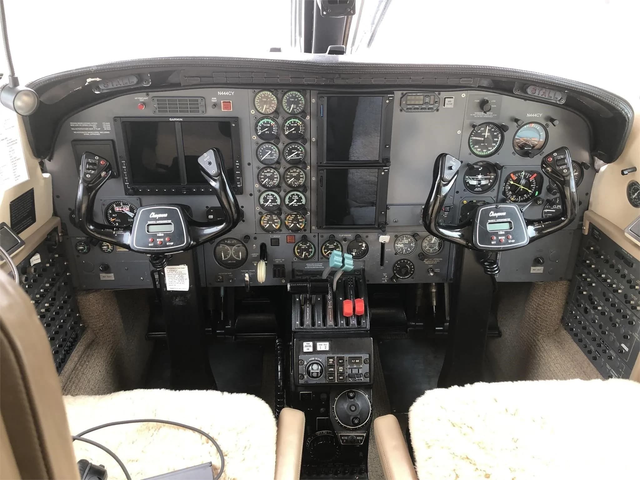 A wide view of a twin-engine aircraft cockpit, showing the instrument panel with multiple displays and gauges, two control yokes, and sheepskin seat covers.