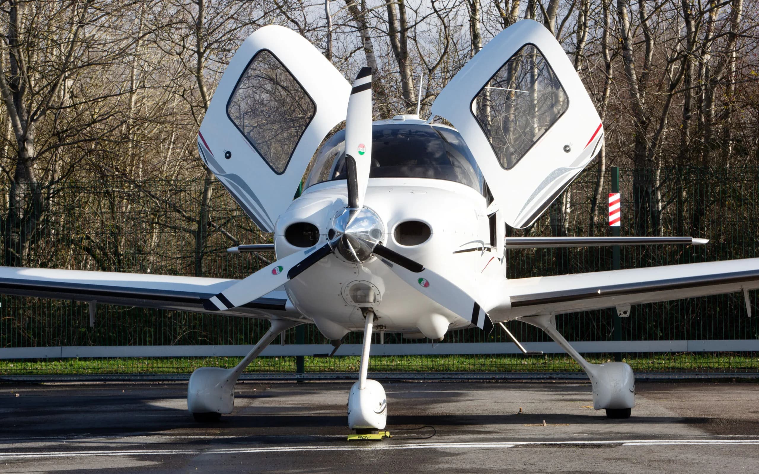 A white Cirrus SR22 G3 aircraft with red and black stripes, registration G-FELC, parked head-on on a tarmac with its gull-wing doors open.