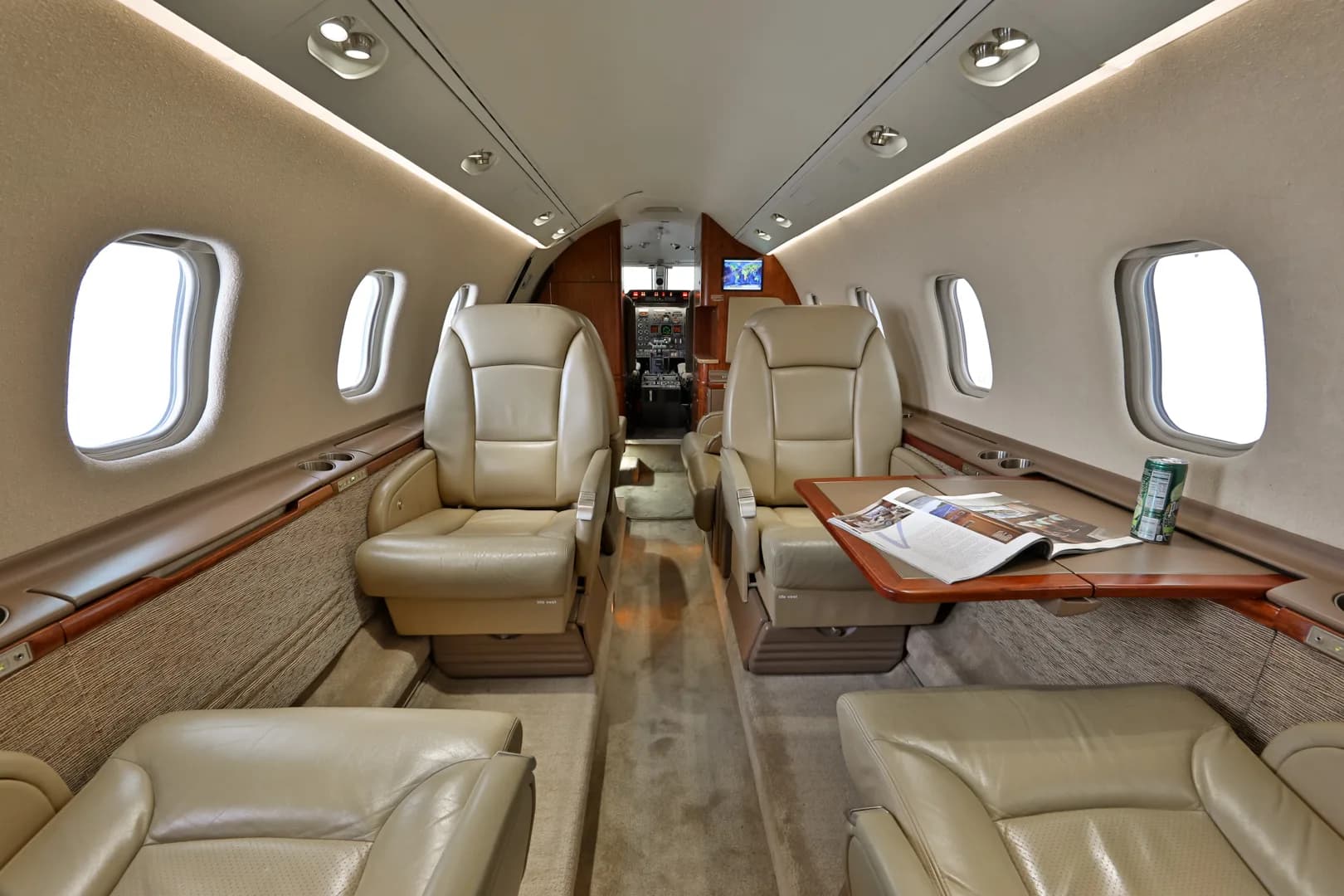The luxurious interior cabin of a business jet with beige leather seats, a fold-out table, and large windows.