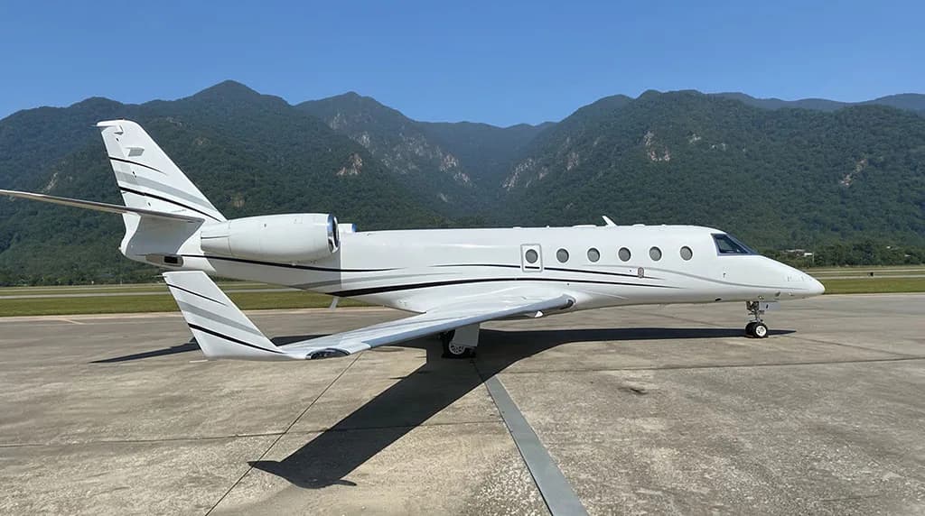 A white business jet with black and grey stripes is parked on an tarmac, viewed from the side.