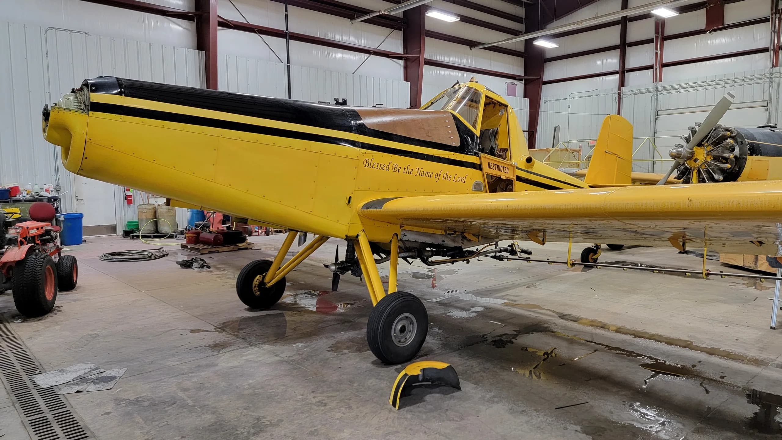 A yellow and black crop duster aircraft, N4941X, with 'Blessed be the Name of the Lord' and 'RESTRICTED' text, parked inside a hangar.