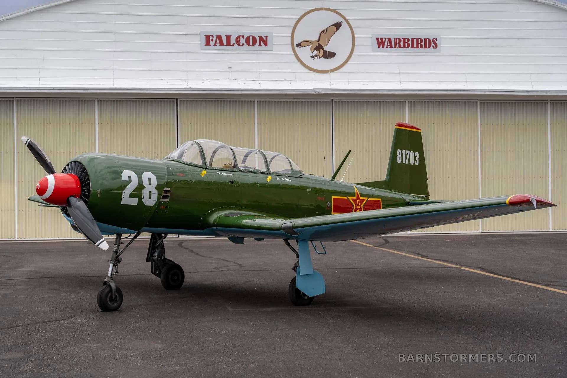 A green Nanchang CJ-6 aircraft with red nose and tail, number 28, parked in front of a hangar with 'FALCON WARBIRDS' logo.