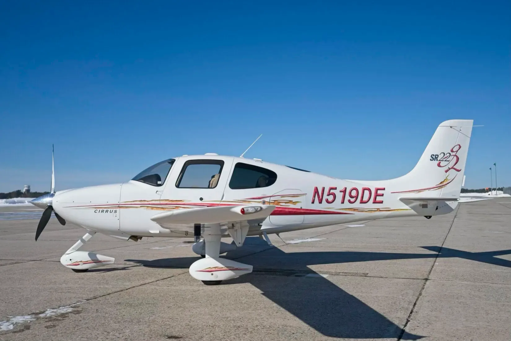 A white Cirrus SR22 aircraft with red and gold stripes, registration N519DE, parked on an airfield under a clear blue sky.