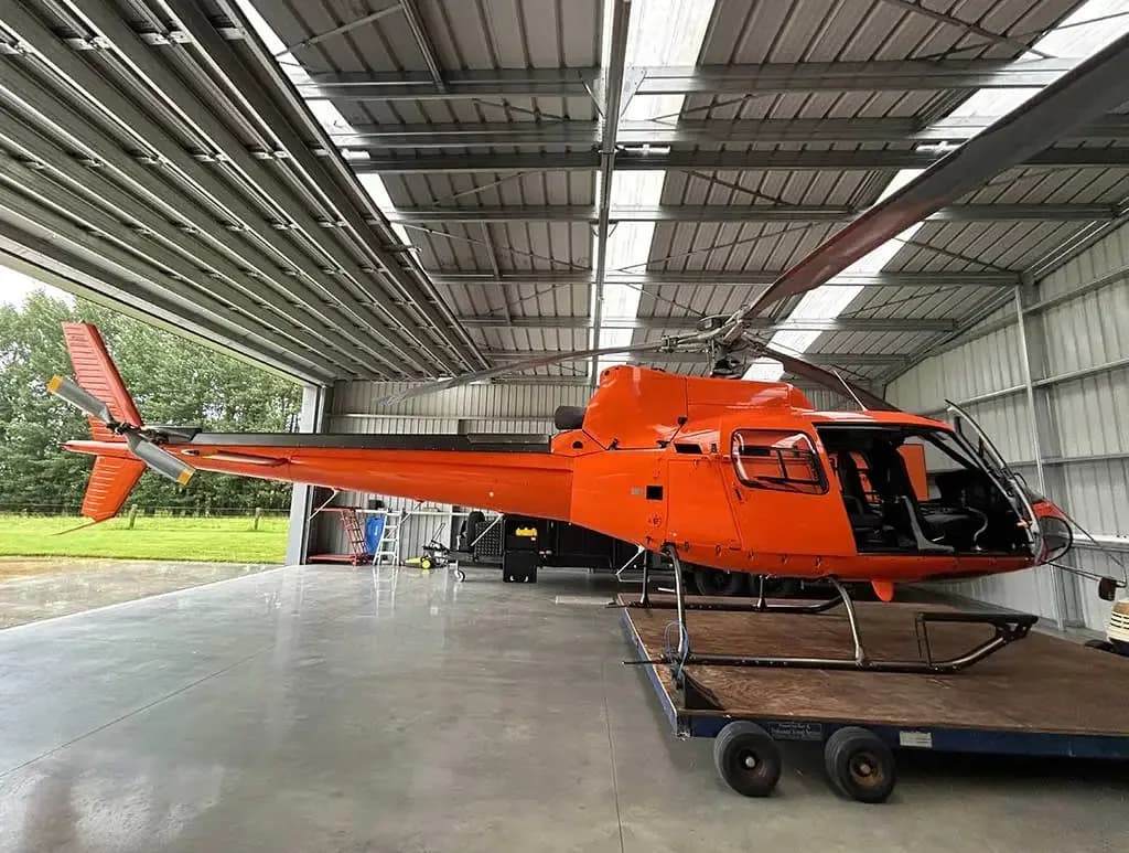 A bright orange helicopter with its main rotor blades extended, parked inside a metal hangar.
