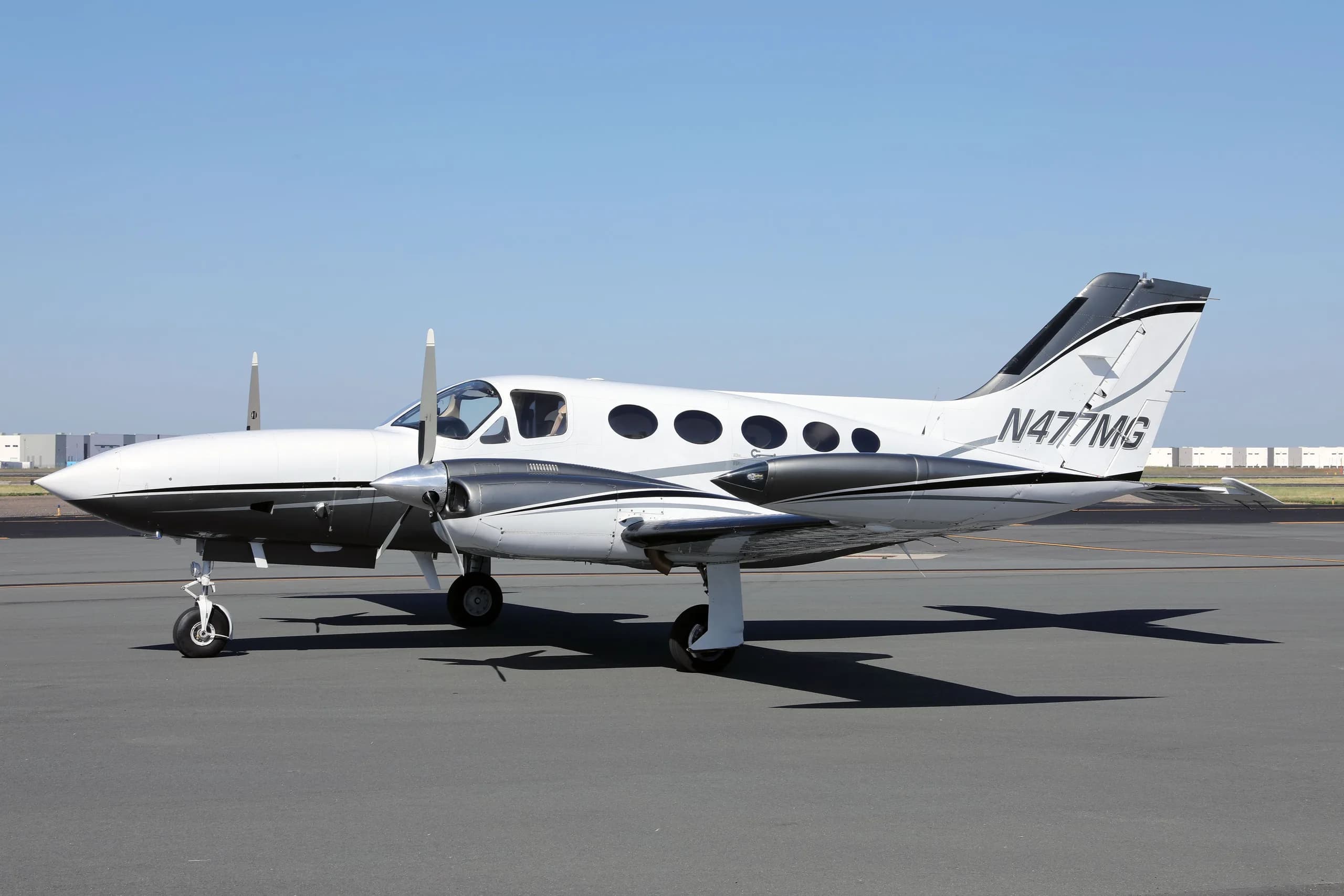 A white and grey twin-engine Cessna 414 Chancellor aircraft, N477MG, parked on an asphalt tarmac under a clear sky.