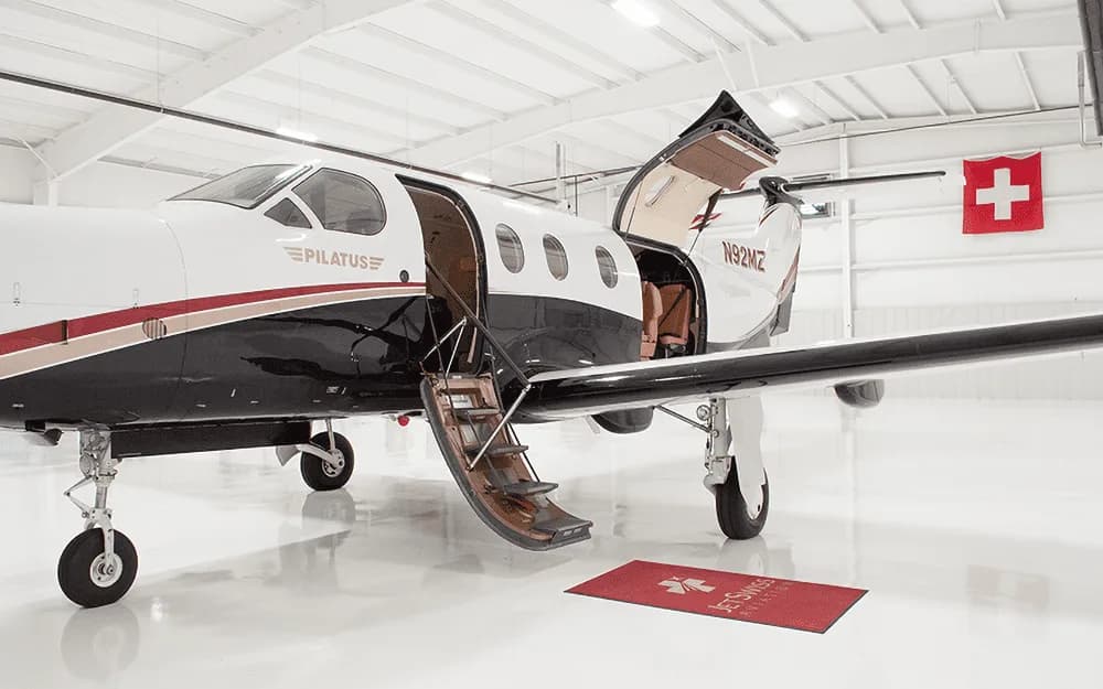 A white and black Pilatus PC-12 aircraft with tail number N92MZ is parked in a hangar with its main entry door open and stairs extended.