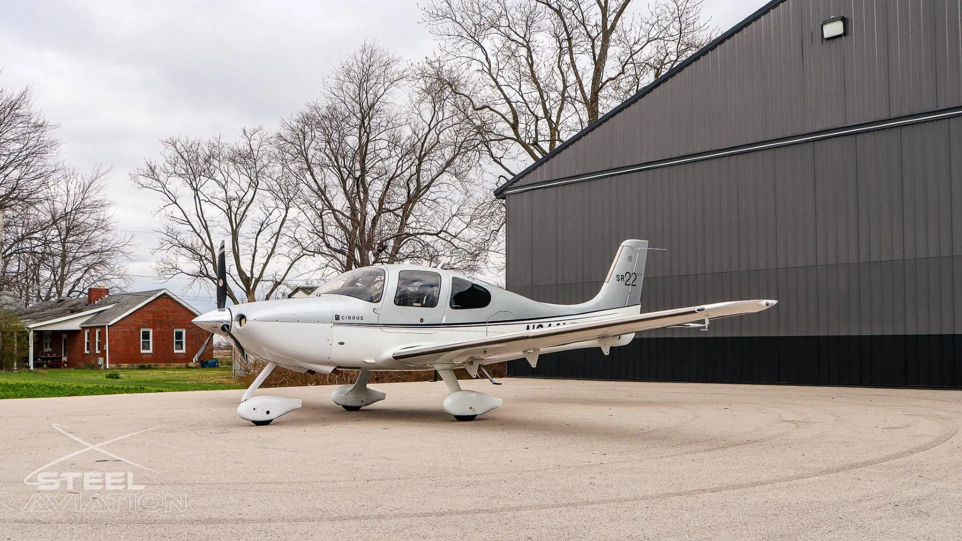 A side view of a white and silver Cirrus SR22 aircraft (N814LT) parked on an apron with trees and a house in the background.