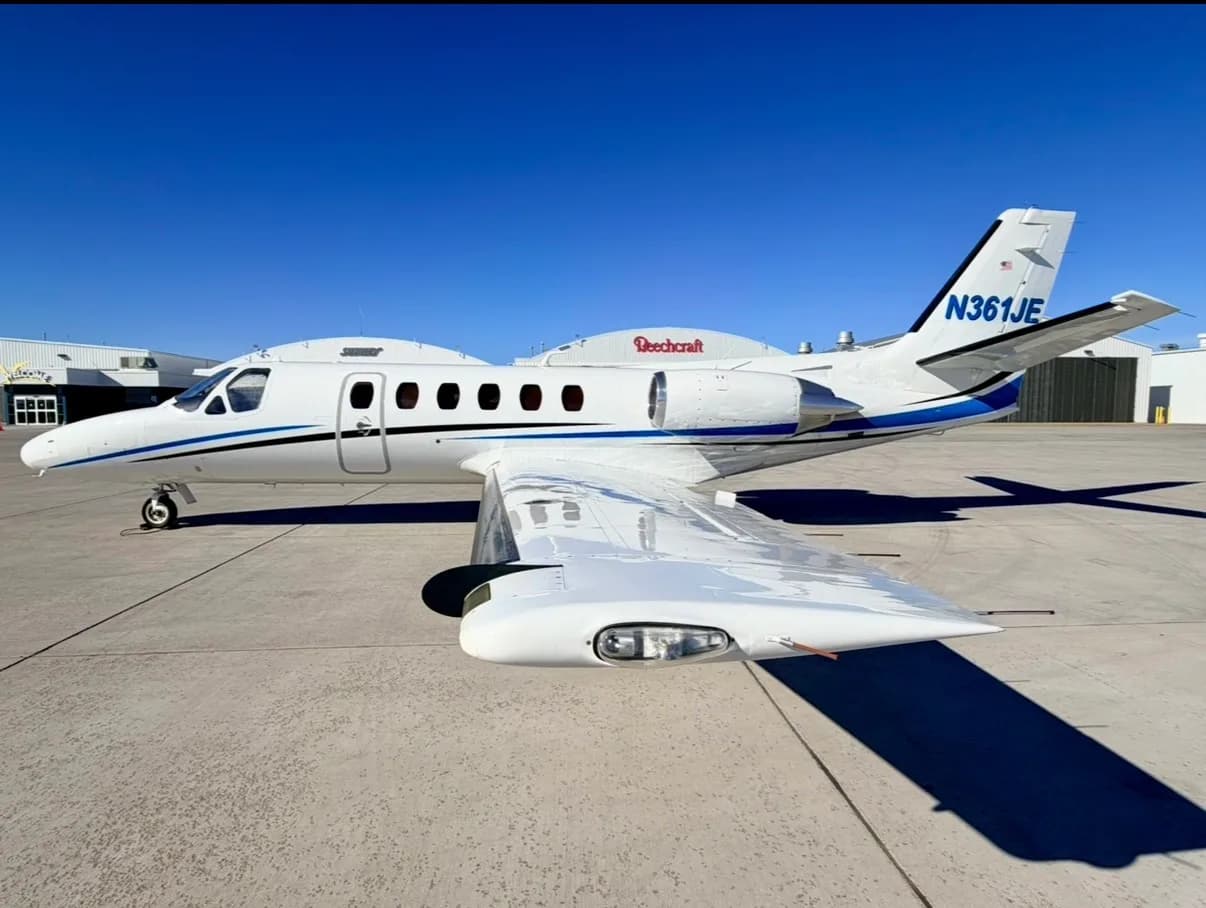 Side view of a white Cessna CitationJet with blue and black stripes, registration N361JE, parked on an airfield under a clear blue sky.
