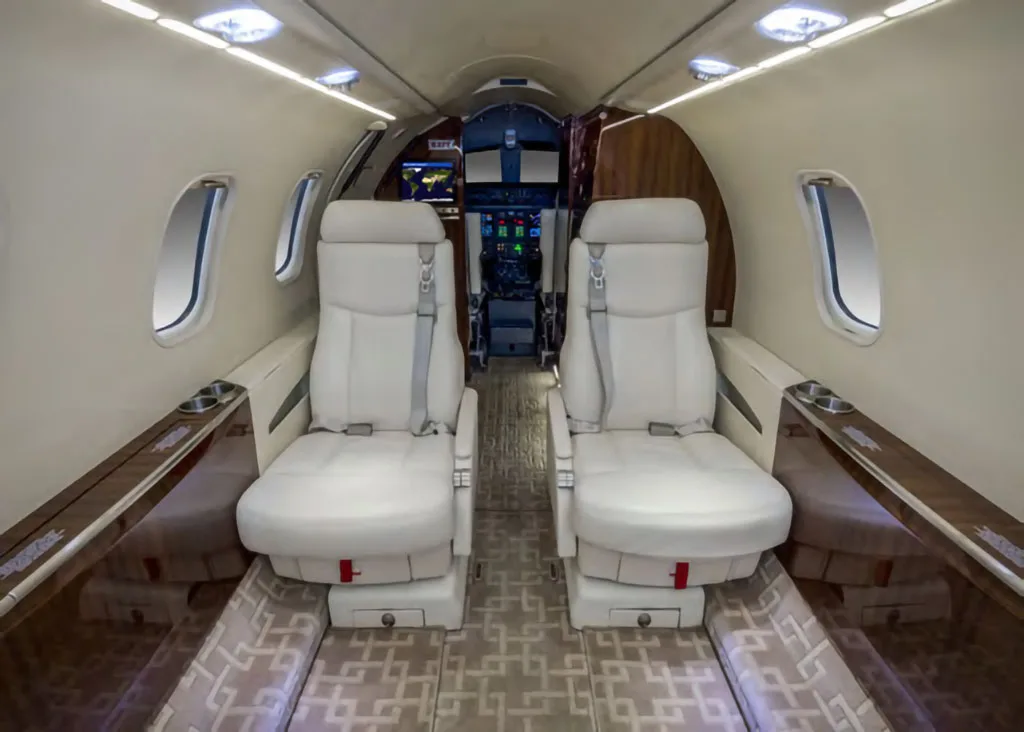 View from the rear of a private jet cabin, showing light-colored seats and the entrance to the cockpit with screens visible.