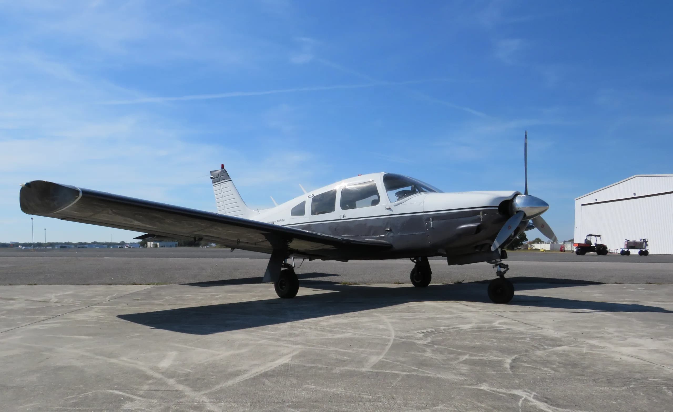 A white and grey Piper Cherokee Arrow single-engine aircraft is parked on an airfield under a clear blue sky.
