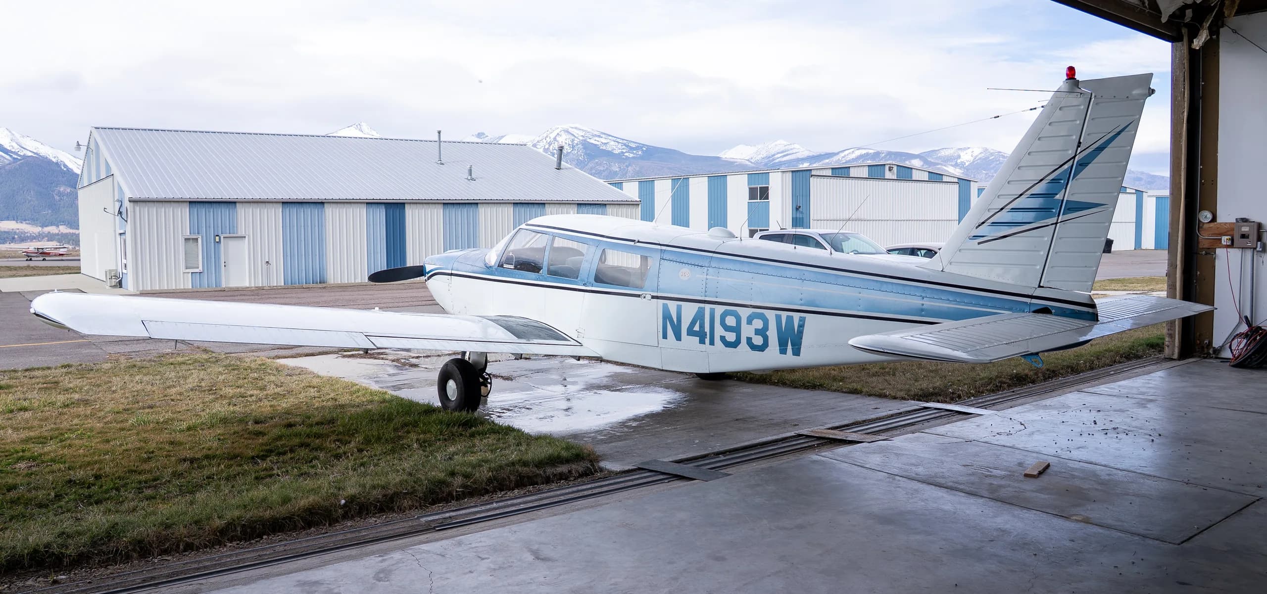 Side view of a white and blue single-engine propeller aircraft N4193W parked outside a hangar with snowy mountains in the background.