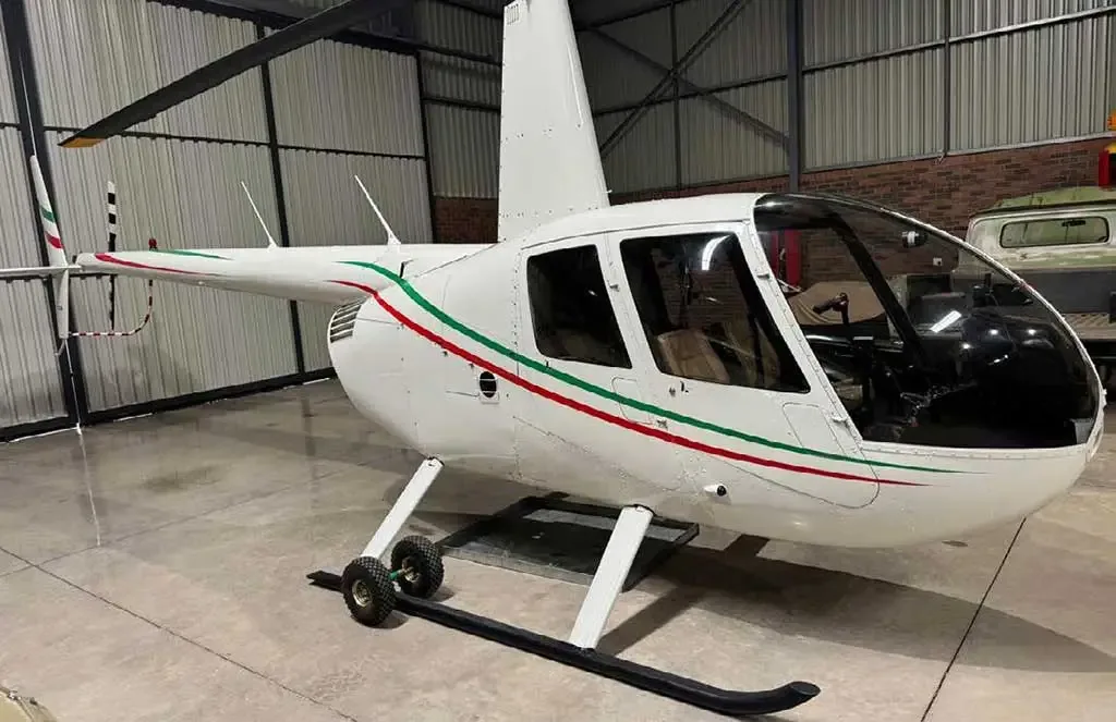 A white Robinson R44 helicopter with green and red stripes is parked inside a hangar, viewed from the front-right showing the right skid.