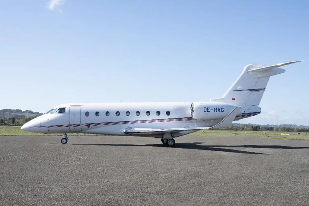 A white business jet with blue and red stripes, seen from the side, parked on an asphalt tarmac.