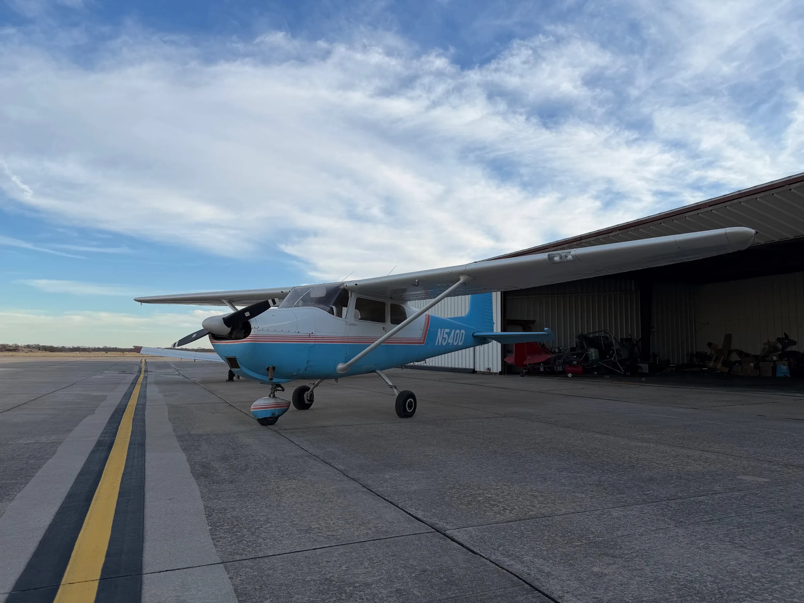 A front-left view of a white and light blue Cessna 172, registration N540D, parked on an airfield with hangars.