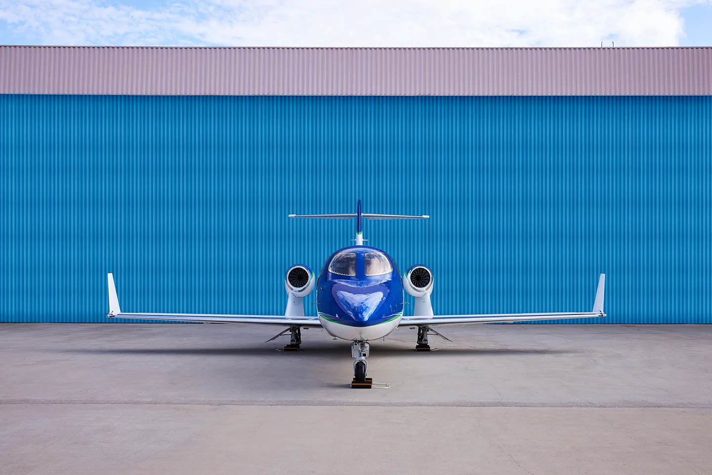 Front view of a white and blue HondaJet Elite with a green stripe, parked on an airfield in front of a blue hangar.
