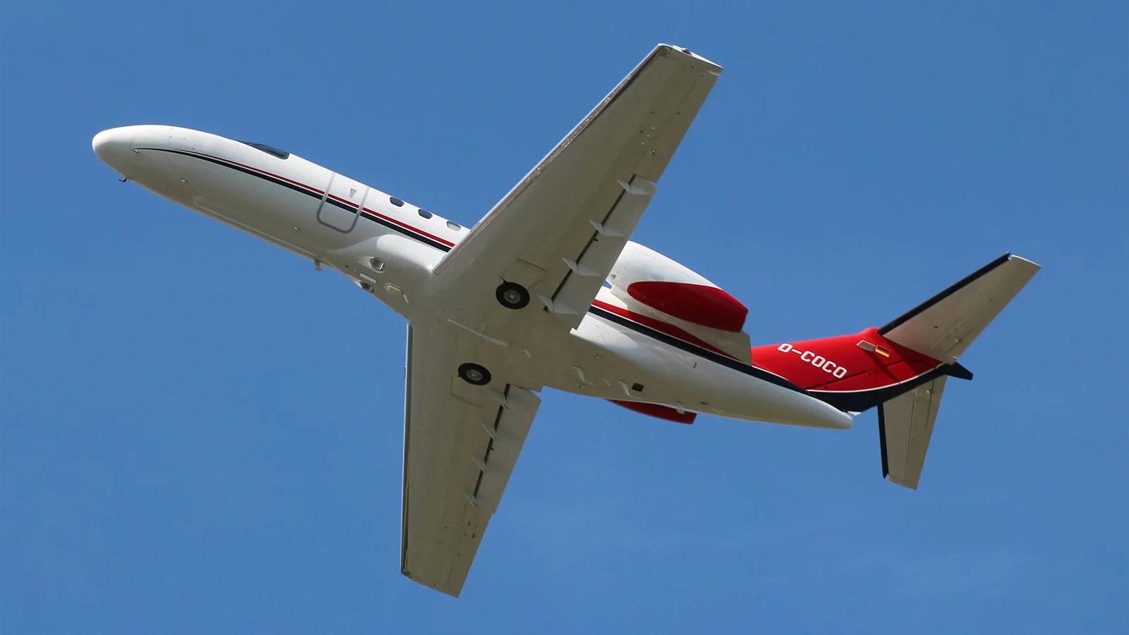 A white Citation CJ4 business jet with red and black stripes and German registration D-COCO in flight, viewed from below.