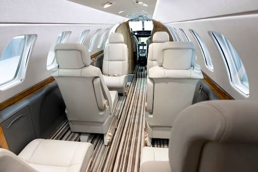 A view down the aisle of the Cessna Citation CJ3+ cabin, showing multiple rows of light cream leather seats and a striped floor.