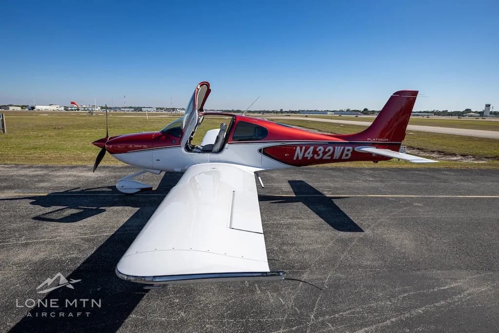 An overhead front-side view of the red and white Cirrus SR20 aircraft (N432WB) with its doors open, parked on an airport tarmac.