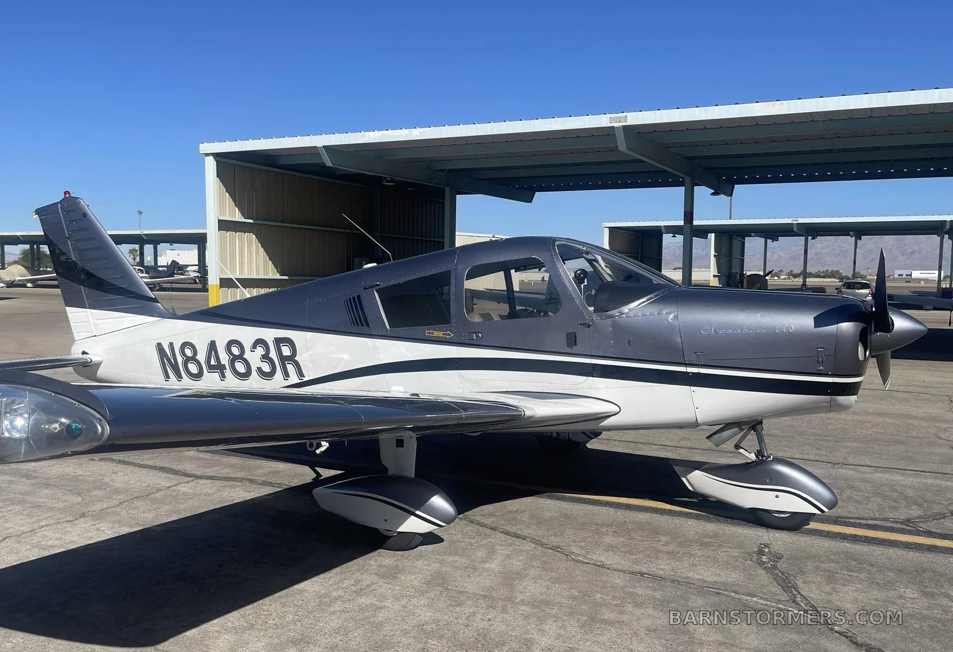 A side view of a Piper Cherokee 140 aircraft in dark gray and white, parked on an airfield under a clear blue sky.