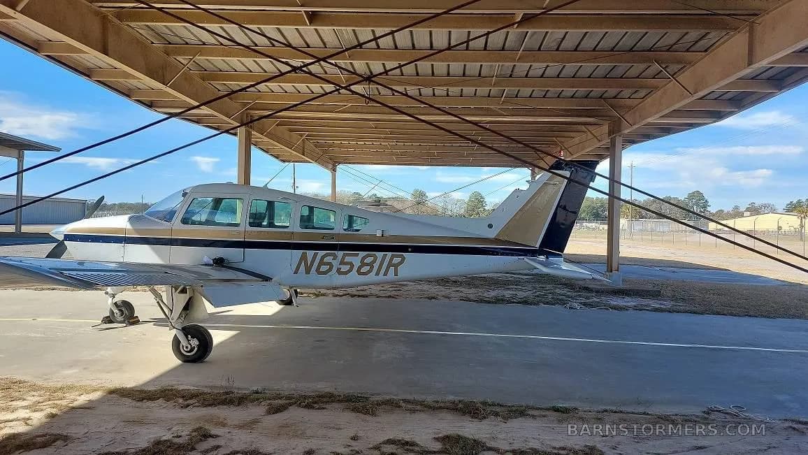A light-colored single-engine aircraft with black and gold stripes, tail number N6581R, parked under a metal canopy.