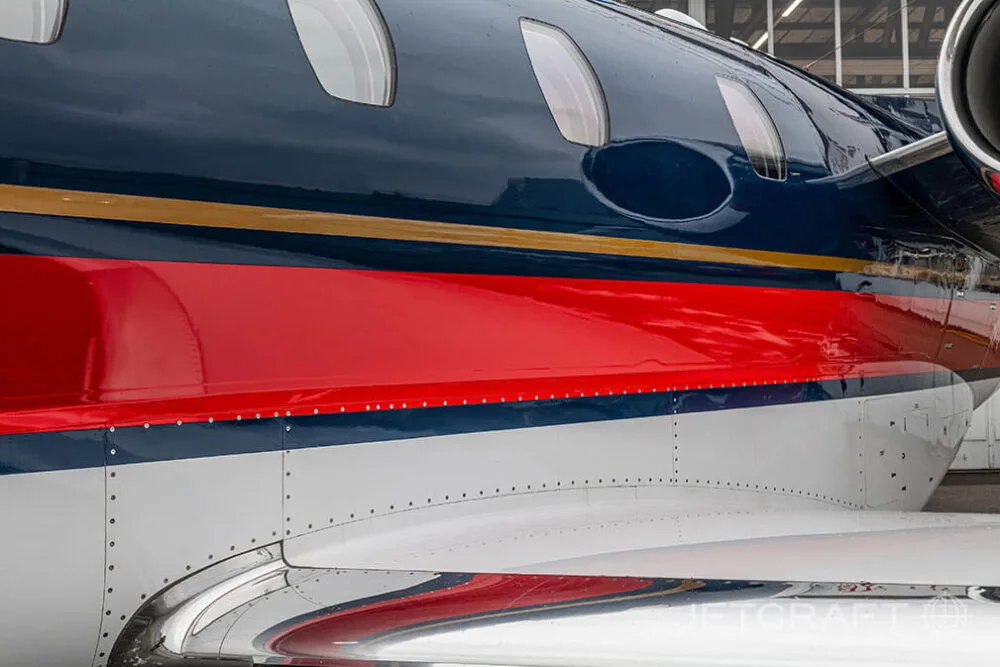 A close-up view of the dark blue, red, and white fuselage and polished wing root of a business jet.