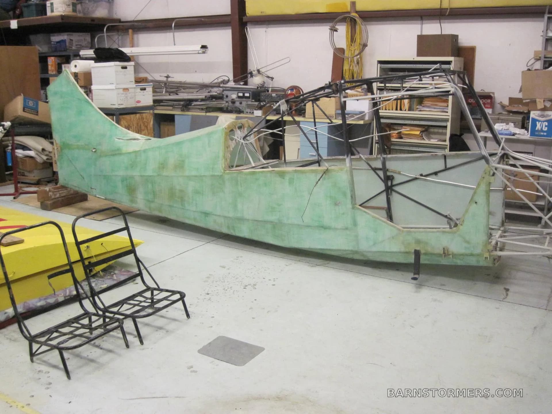 A light green aircraft fuselage frame and skin under construction in a workshop, with two metal seats in the foreground.