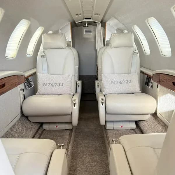 Interior view of a private jet cabin with light-colored leather seats and pillows embroidered with 'N747JJ'.