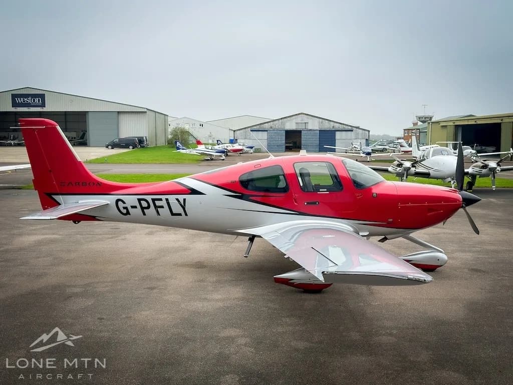 A red and white Cirrus SR22 aircraft (G-PFLY) parked on a tarmac with other aircraft and hangars in the background.