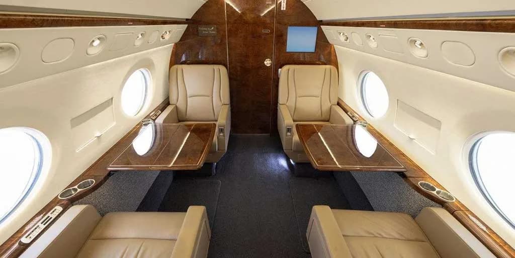 The elegant cabin interior of a business jet with tan leather seats, wood tables, and overhead lighting.