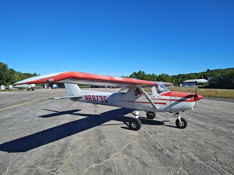 A white and red Cessna 150/152 aircraft with registration N6973G is parked on an asphalt tarmac under a clear blue sky.