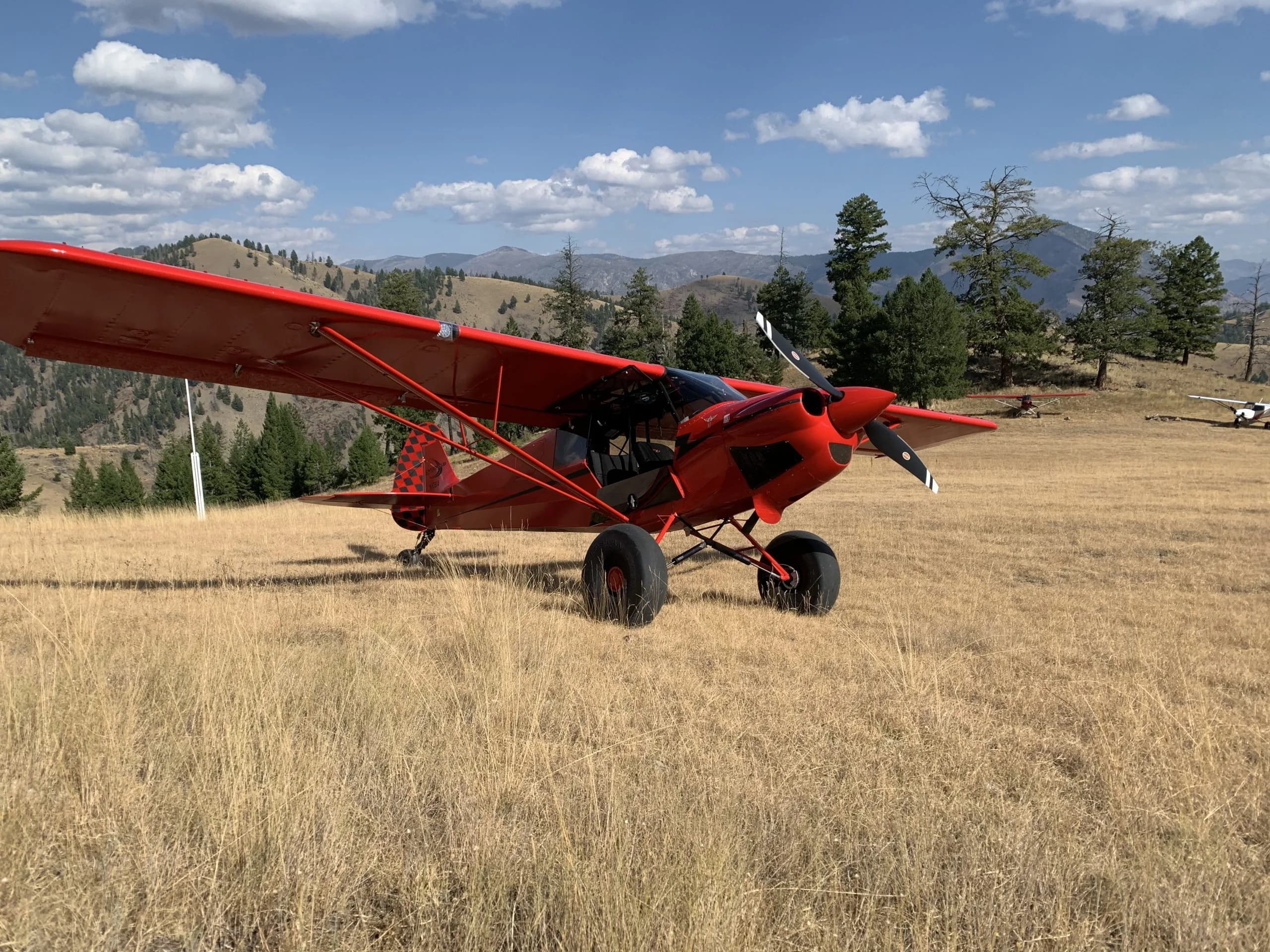 A red bush plane with large tundra tires and a checkered tail is parked on a grassy mountain airstrip under a blue sky.