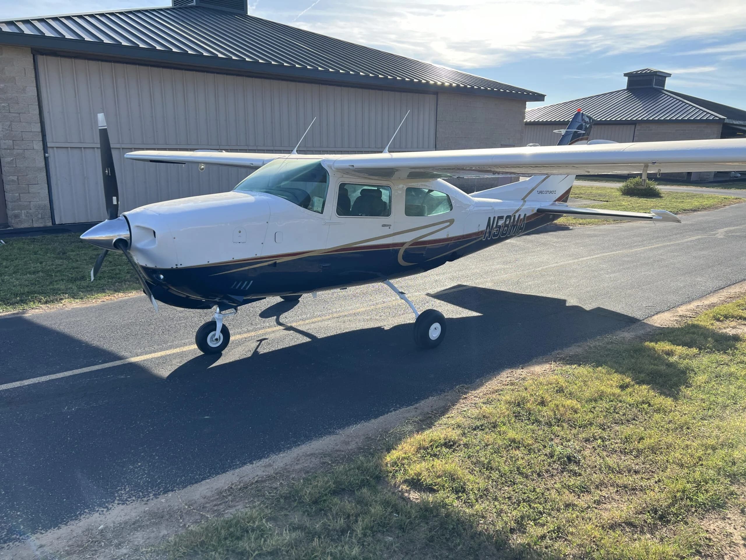 A white and blue Cessna 210, N58MA, with gold trim, parked on an asphalt taxiway with hangars in the background.