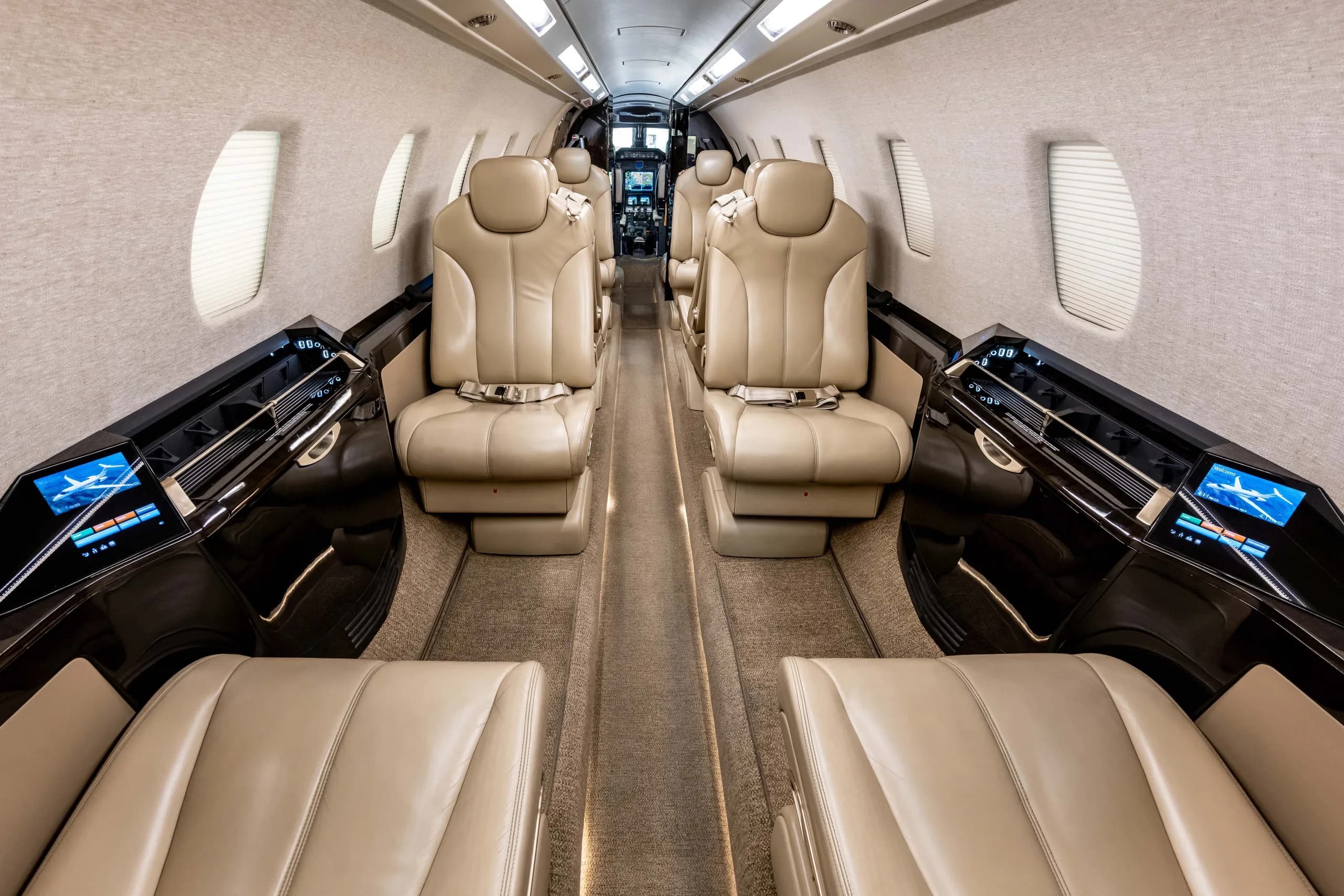 The interior cabin of a business jet with light tan leather seats and dark wood accents, looking forward towards the cockpit.