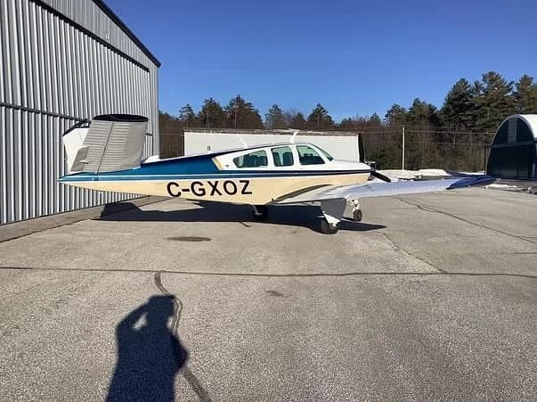 A beige and blue single-engine aircraft with registration C-GXOZ is parked next to a metal hangar on a sunny day.