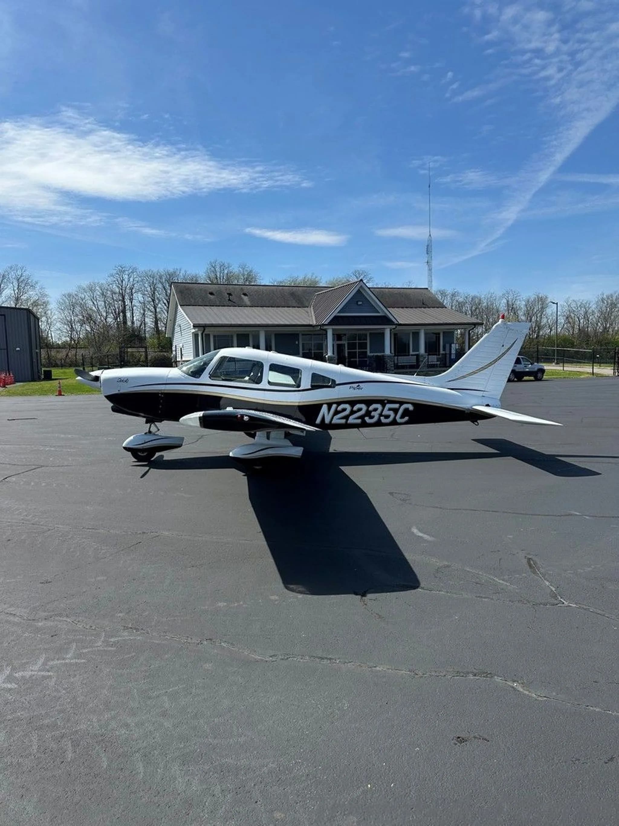 A white and black Piper aircraft with gold trim, registration N2235C, parked on an asphalt ramp with a building in the background.