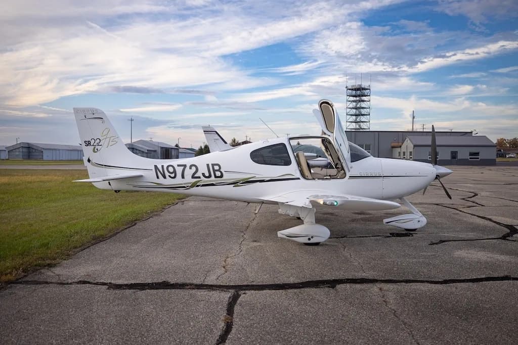 A white Cirrus SR22T aircraft, N972JB, is parked on an airfield with its doors open, viewed from the right side.