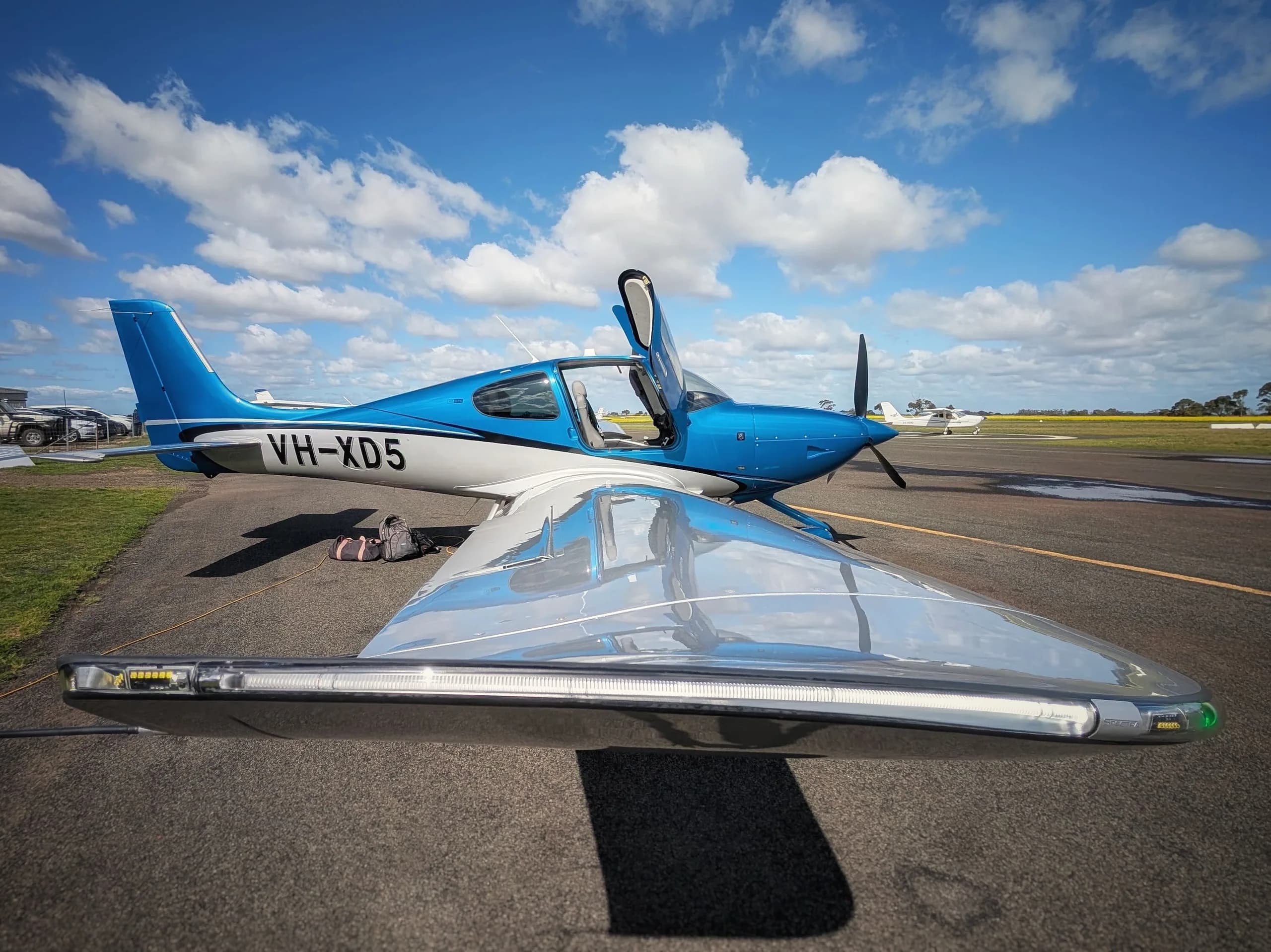 A blue and white single-engine Cirrus SR22 aircraft with registration VH-XD5 parked on an airfield with its doors open.