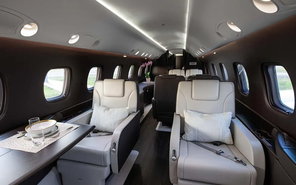 The elegant interior of a private jet cabin, with light leather seats, a dining table, and overhead lighting.