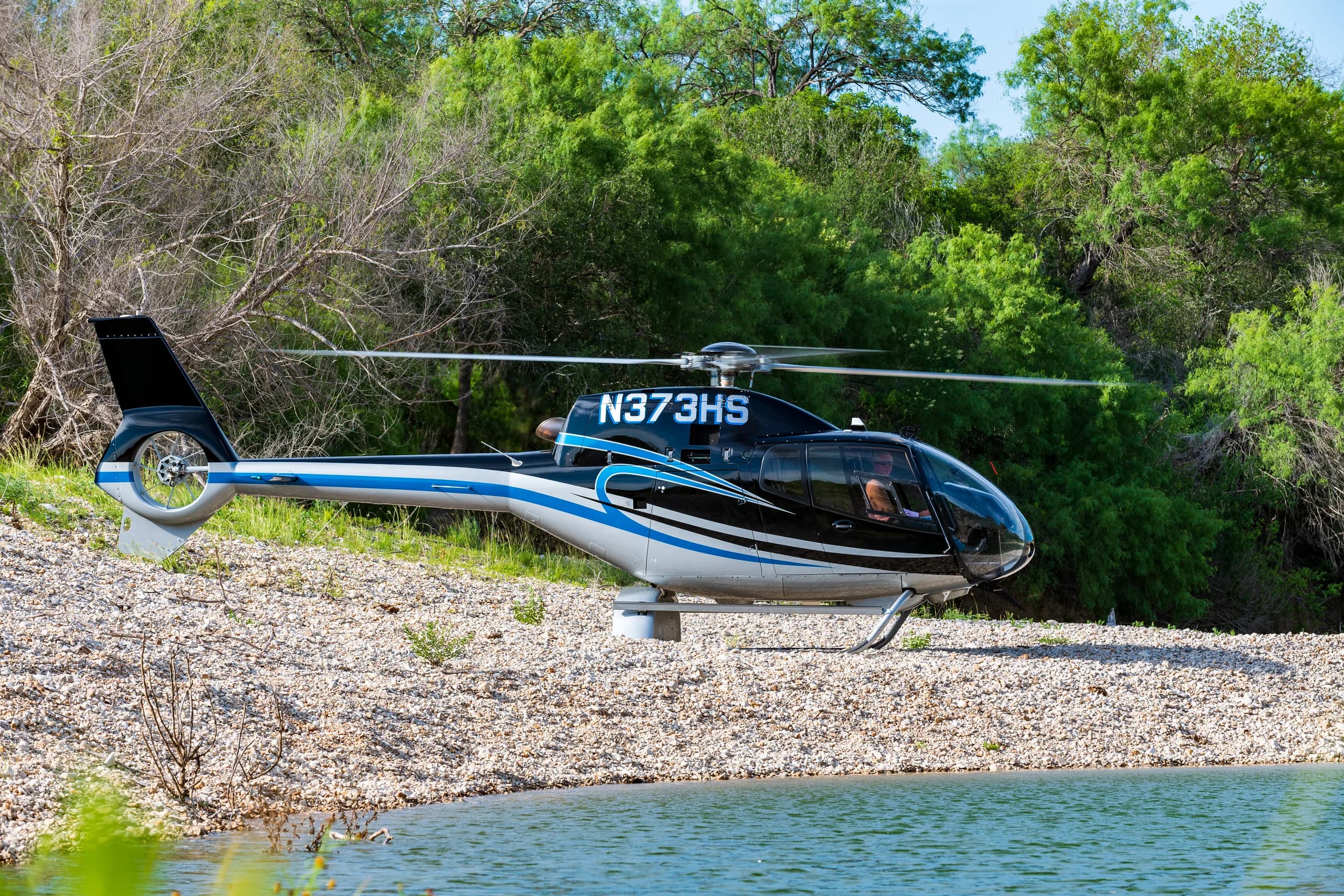 A black and blue helicopter (N373HS) hovers just above the water next to a rocky shore.