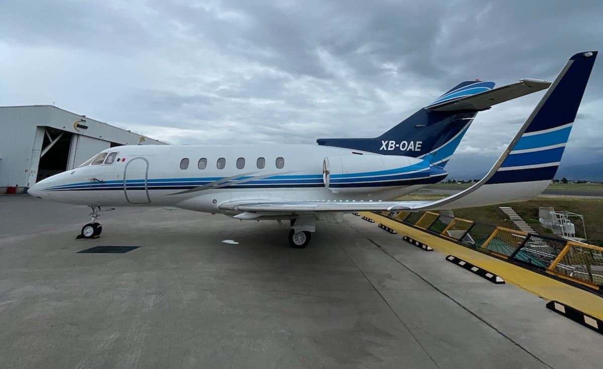 A white business jet with blue stripes (XB-OAE) parked on a tarmac next to a hangar.