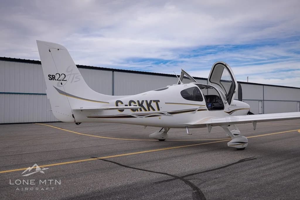 A rear three-quarter view of a white Cirrus SR22 aircraft with registration C-GKKT, doors open, on an airfield.