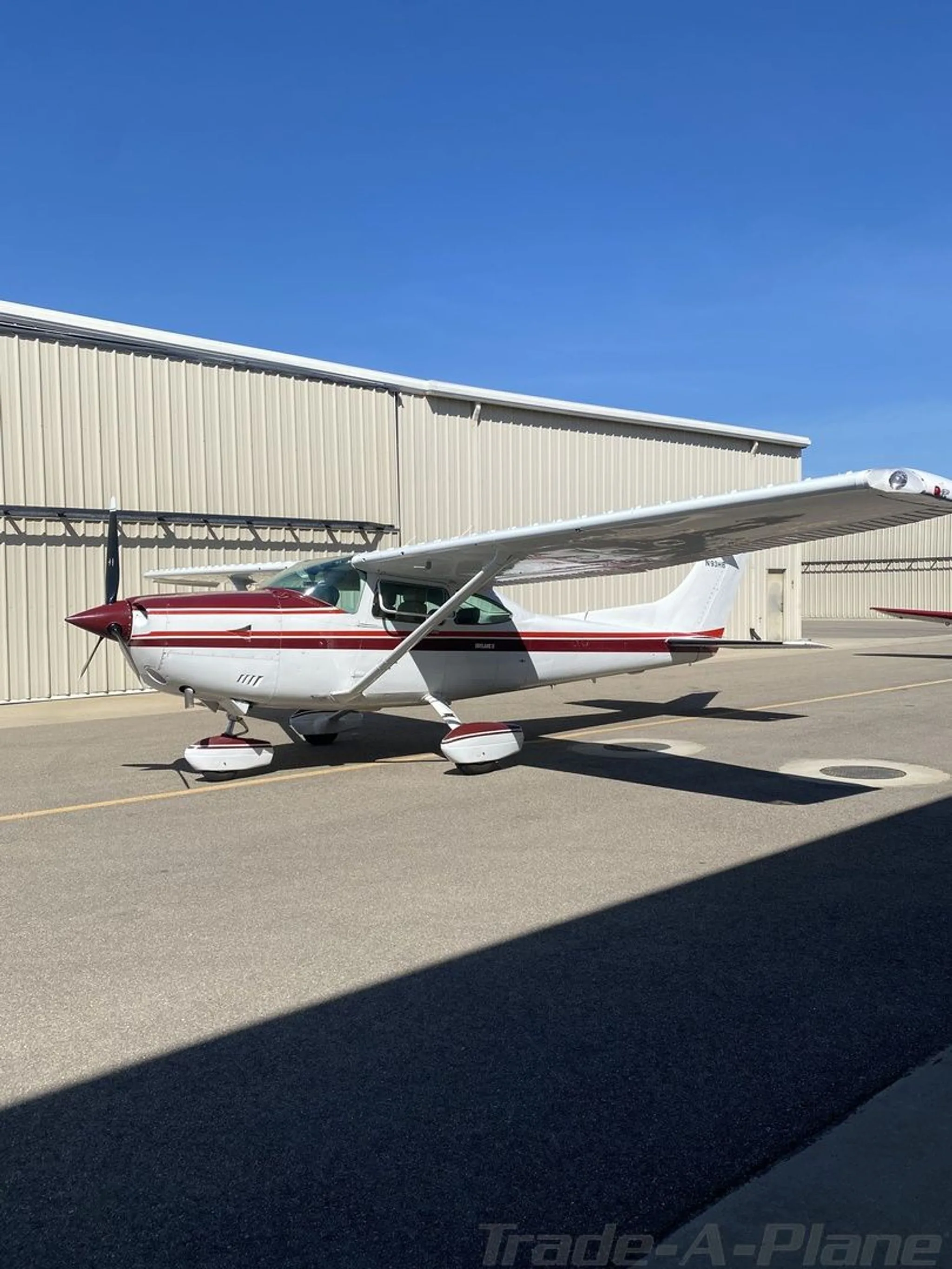 A white Cessna 182 with a red stripe is parked on an asphalt ramp next to a hangar under a clear blue sky.