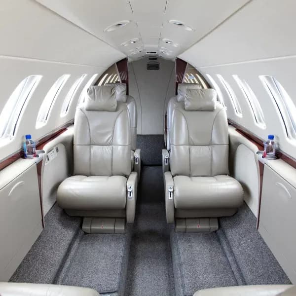 The aircraft cabin interior features two rows of beige leather club seats with water bottles on side tables.