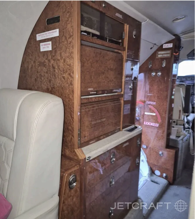 A wood-paneled galley or storage unit inside a private jet cabin, with various compartments and a light-colored seat in the foreground.